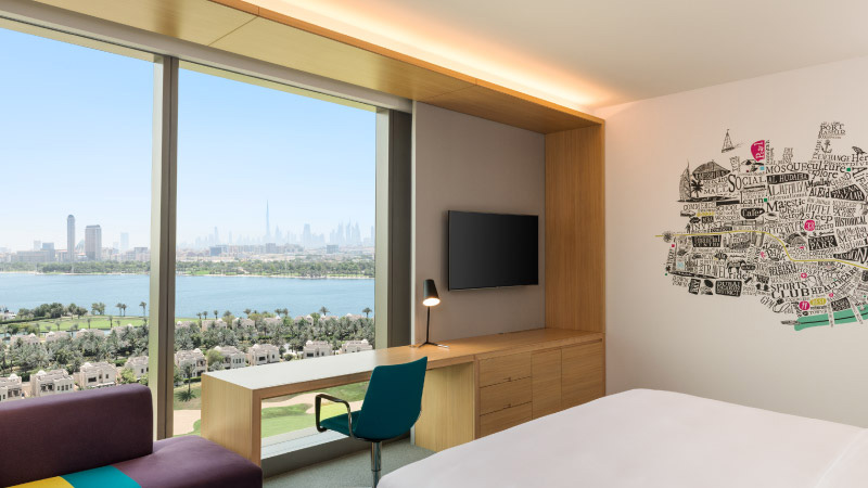Modern hotel room with a large window offering a city skyline and water view. Features minimalistic decor, a desk, chair, and artistic wall design.
