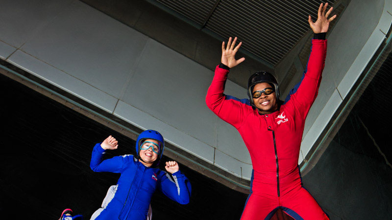 Two people in flight suits enjoy indoor skydiving. One wears a blue suit, the other red. Both are smiling, conveying excitement and fun.