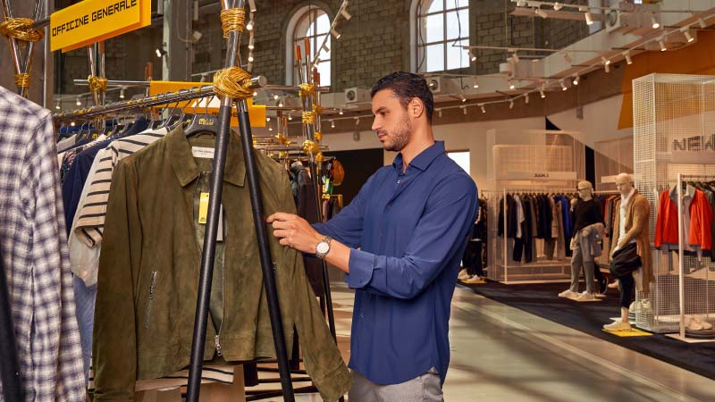 A man in a blue shirt browses a clothing rack in a stylish store, focused on a green suede jacket. The setting is modern and well-lit, conveying a casual shopping experience.