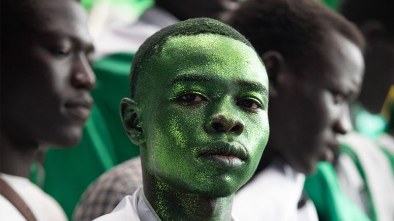 A young person with green glitter covering their face stands in focus, surrounded by others in soft focus. The scene conveys a festive and spirited atmosphere.