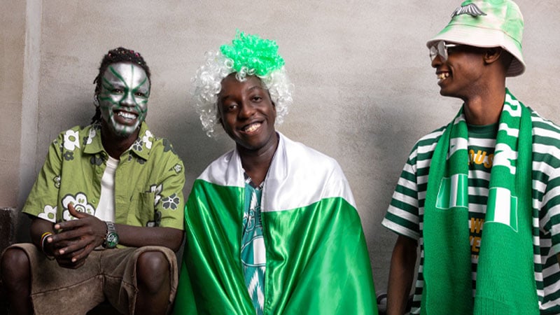 Three men dressed in green and white sit against a gray wall, smiling. One has face paint, another wears a wig and flag, and the third sports a hat. They convey joy and camaraderie.