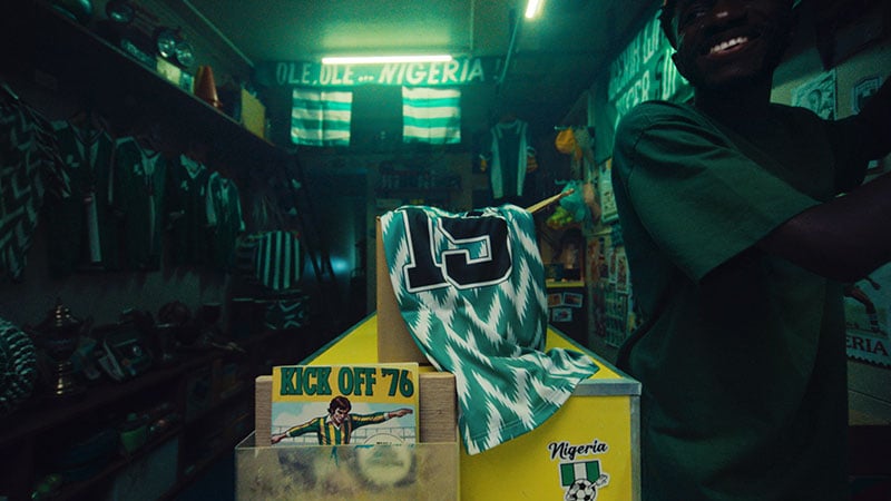 Dimly lit room with a green glow, filled with Nigeria football memorabilia. A jersey with number 15 draped on a stand. Person smiling on the right.