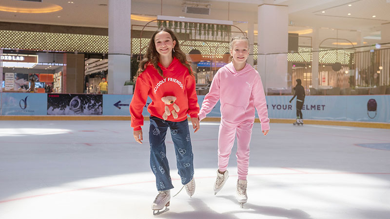 Two girls wearing colorful sweaters ice skate together indoors, holding hands and smiling, with a shopping mall visible in the background.